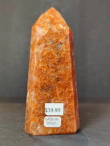 Polished sunstone obelisk tower with orange and reddish hues, standing upright against a neutral gray background with price and origin labels visible.