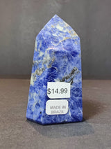 Polished sodalite obelisk sculpture in deep blue with white and gray marbled patterns, standing upright on a dark surface with price and origin labels.