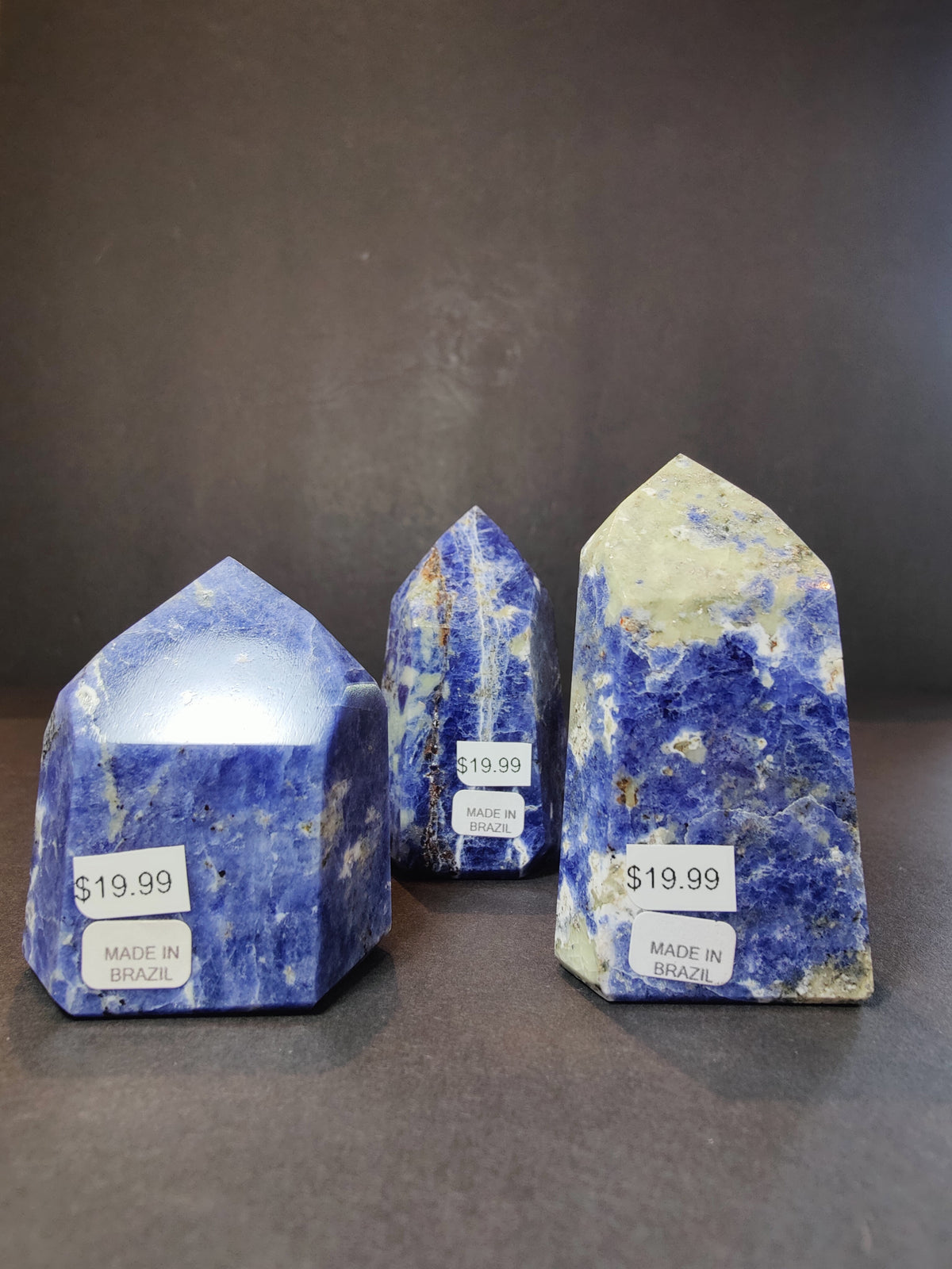 Three sodalite polished obelisk towers in blue with white and black veining, each with a $19.99 price sticker and 'Made in Brazil' label, shown on a dark surface.