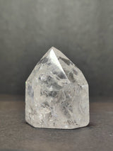 Clear quartz crystal point with internal crackle patterns, standing upright on a dark surface against a blurred gray background.