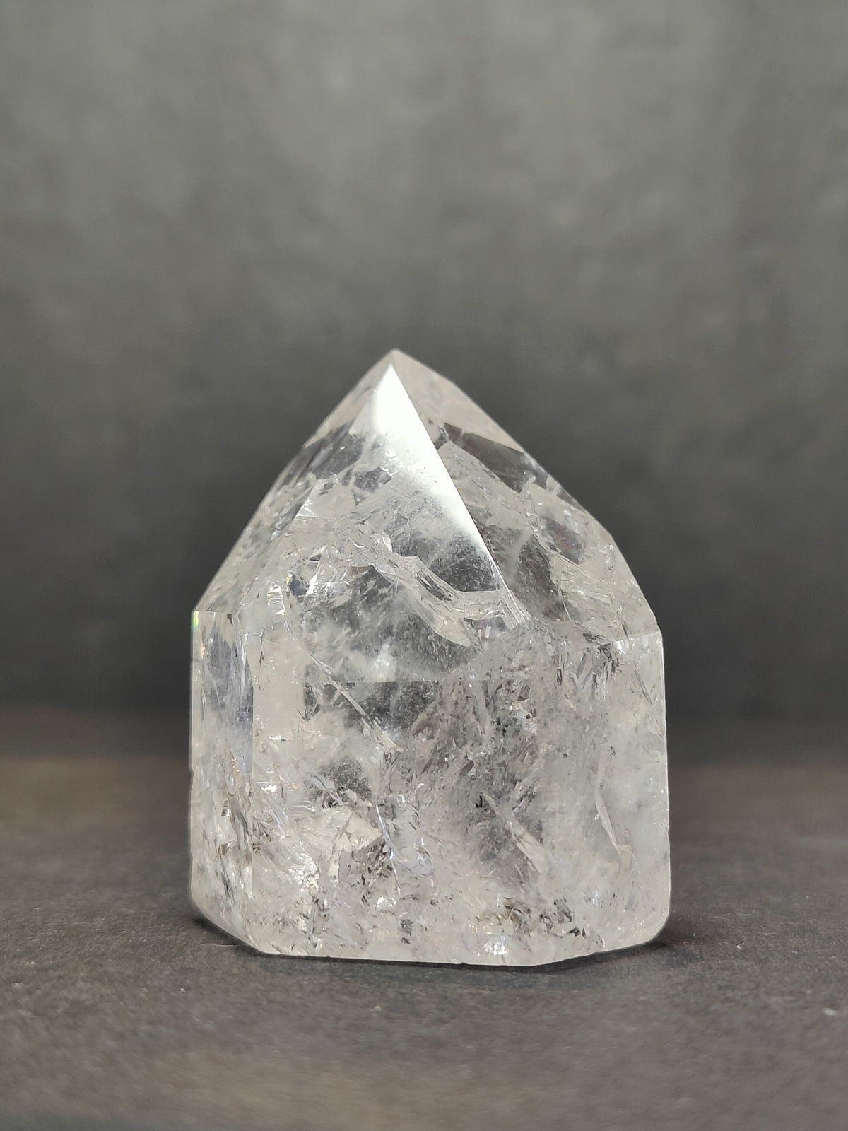 Clear quartz crystal point with internal crackle patterns, standing upright on a dark surface against a blurred gray background.