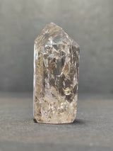 Transparent quartz crystal point with internal crackle patterns, standing upright on a gray surface against a blurred gray background.