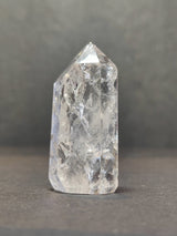 Clear quartz crystal point with internal fractures and inclusions, standing upright on a neutral gray surface against a softly blurred gray background.