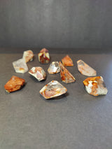 Nine small polished quartz freeform stones with translucent, amber, and reddish inclusions arranged loosely on a dark gray surface against a dark background.