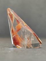Polished translucent quartz freeform with internal reddish-brown and orange amphibole inclusions, shaped with angular facets against a gray background.