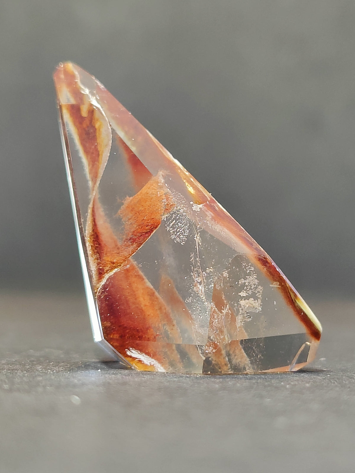 Polished translucent quartz freeform with internal reddish-brown and orange amphibole inclusions, shaped with angular facets against a gray background.