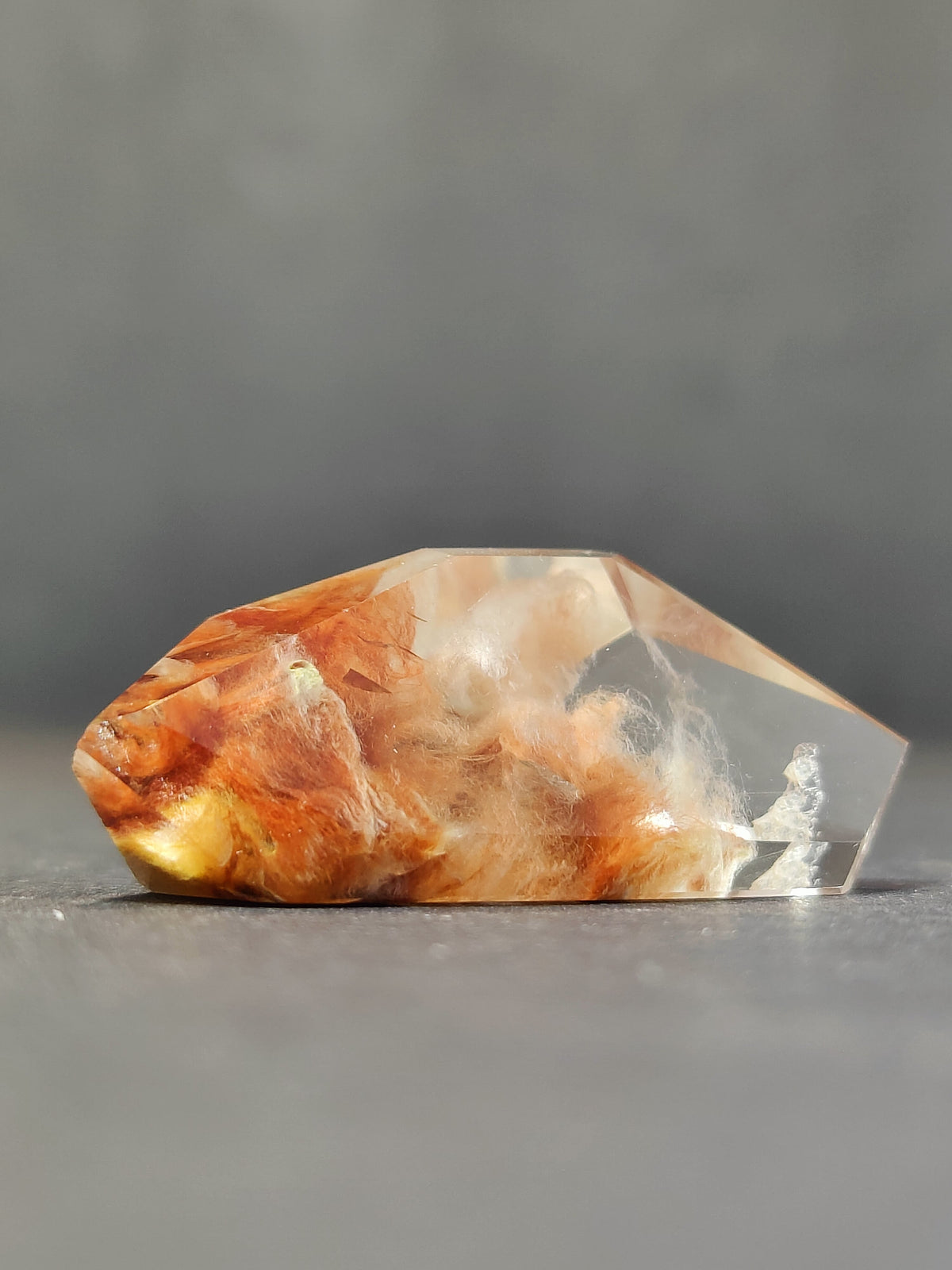 Polished amphibole quartz freeform with translucent sections blending orange, yellow, and smoky inclusions on a gray background.