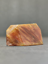 Polished amphibole quartz freeform with amber and reddish-brown striations, displayed against a plain gray backdrop.