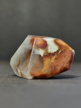 Polished amphibole quartz freeform with a blend of white and rusty orange hues in an irregular faceted shape against a dark gray background.