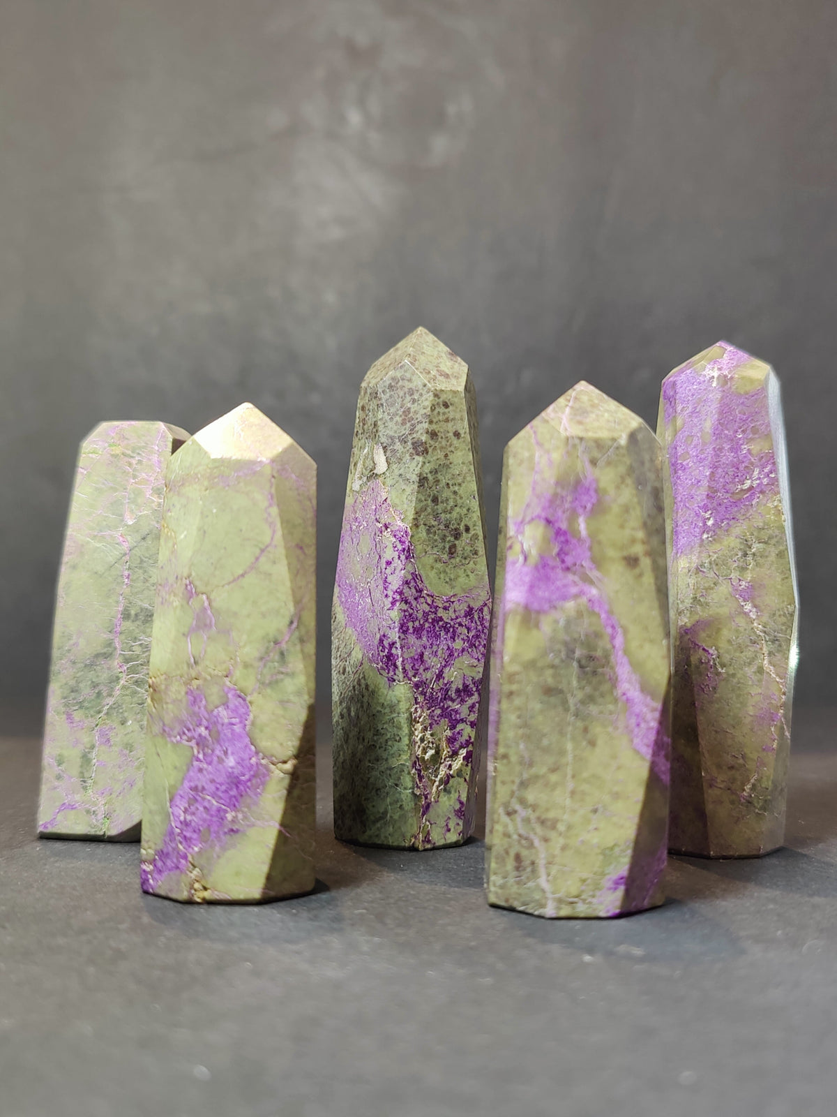 Six polished Atlantisite towers with greenish-gray and vibrant purple marbled patterns standing upright on a dark surface against a dark gray background.