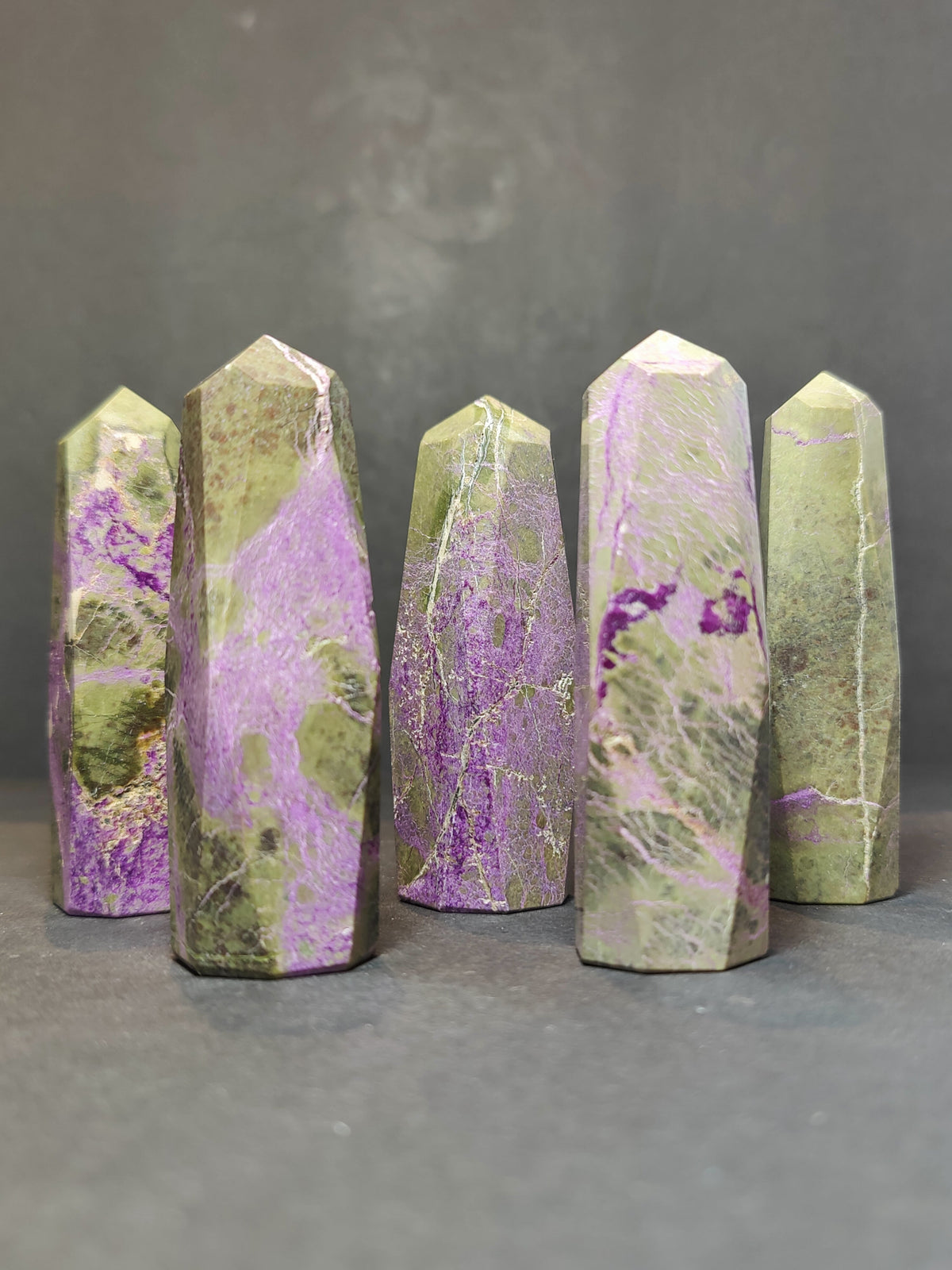 Five polished Atlantisite tower sculptures featuring green and purple marbled patterns, arranged standing upright on a gray surface against a dark gray background.