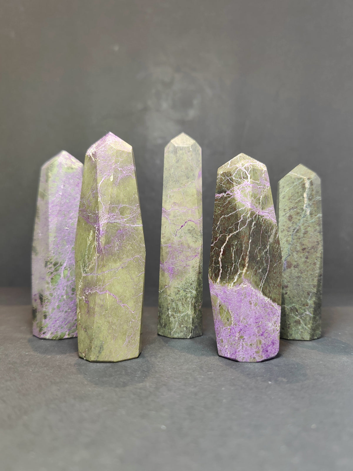Five polished Atlantisite towers with a pointed hexagonal shape, featuring green and purple hues with natural veining, standing upright on a dark surface.
