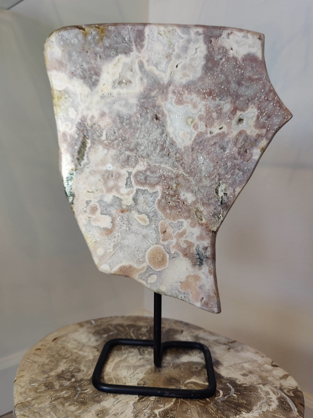Polished pink amethyst slab with intricate white and gray patterns, shaped irregularly and mounted on a minimal black metal stand atop a textured surface.