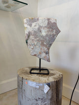 Polished pink amethyst slab with white and gray crystal patterns displayed upright on a black metal stand, placed on a round wooden pedestal against a plain wall.