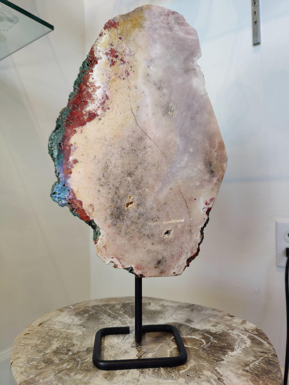 Tall, thin pink amethyst polished slab with beige, red, and green hues on uneven edges, mounted vertically on a black metal stand on a marbled surface.
