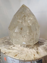 Clear to pale yellow polished natural citrine point with internal fractures and inclusions, standing on a round, textured stone stool with labels and markings.