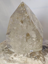 Polished natural citrine crystal point with pale yellow and clear tones displayed on a textured stone surface under soft light.