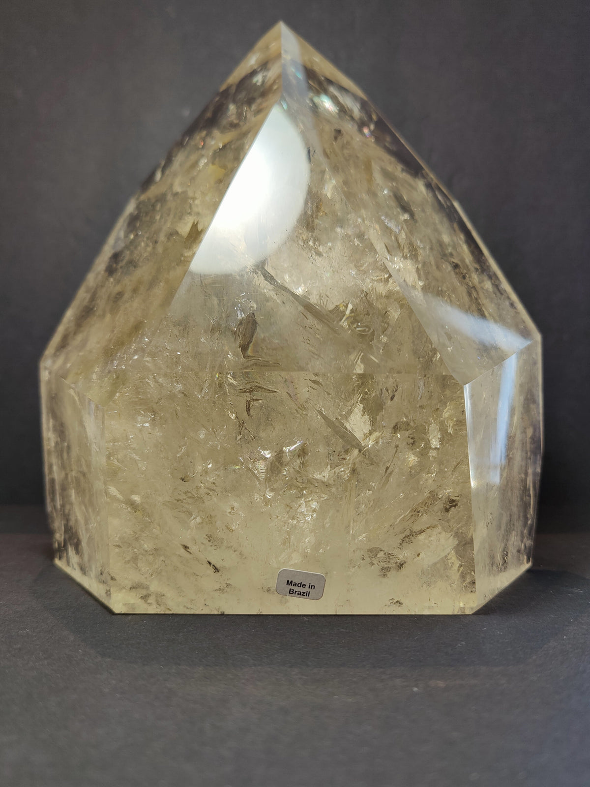 Polished citrine crystal point with a translucent golden-yellow hue, featuring internal fractures and a faceted geometric shape, labeled 'Made in Brazil'.
