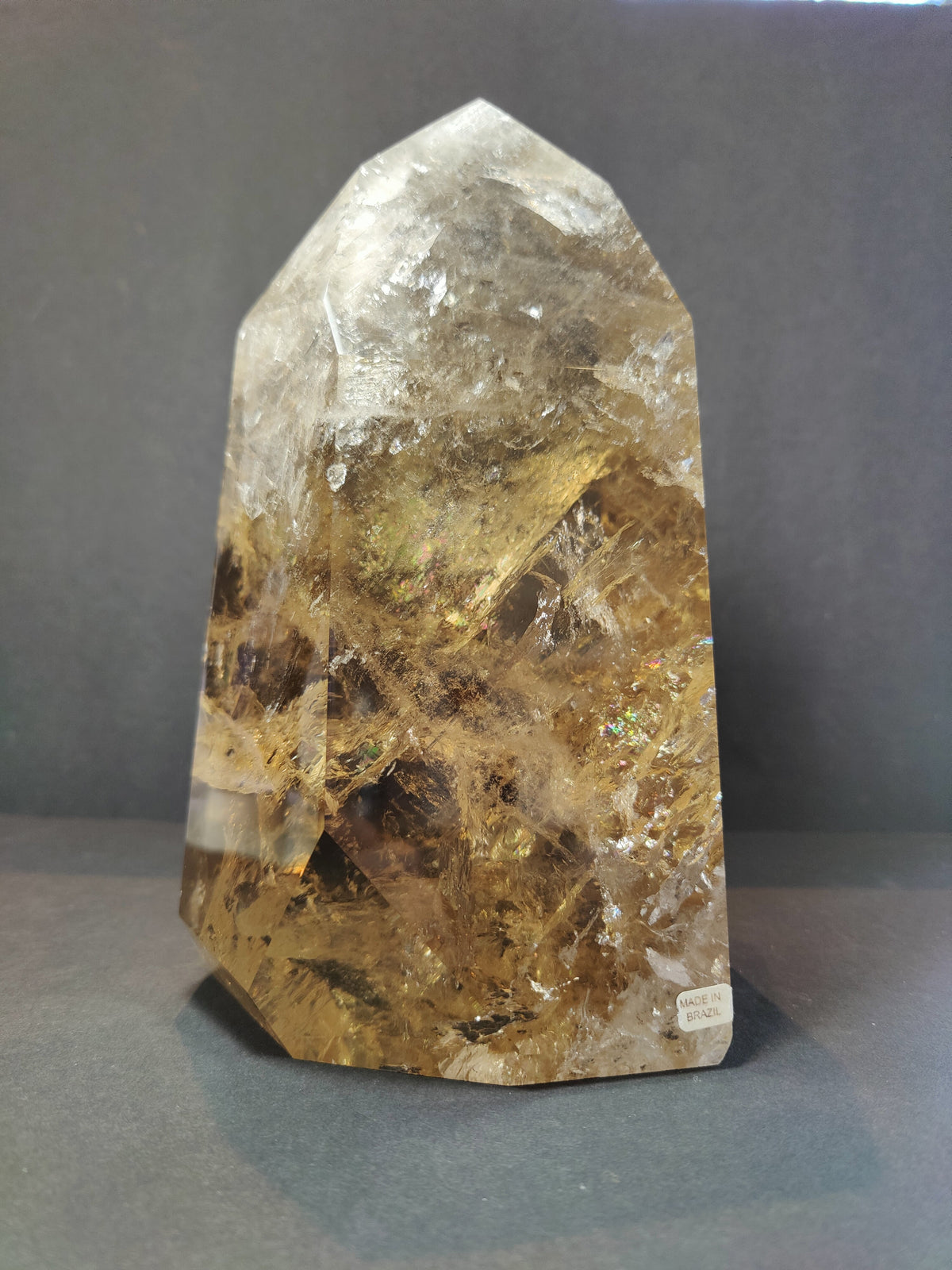 Polished natural citrine point with translucent golden-yellow hues and internal crystal formations, standing upright against a dark grey background.