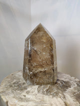 Polished natural citrine crystal point with translucent amber and smoky brown hues, standing upright on a light-colored textured stone surface.