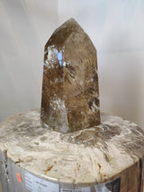 Polished natural citrine crystal point with translucent golden hues and internal fractures, standing vertically on a round, beige patterned stone surface against a light background.