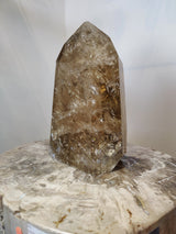 Polished natural citrine crystal point with translucent amber and golden-brown hues, displayed upright on a marbled surface with subtle veining.