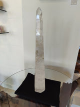 Tall, clear polished quartz obelisk sculpture with natural internal fractures, standing on a black cloth atop a round glass table against a plain white wall.