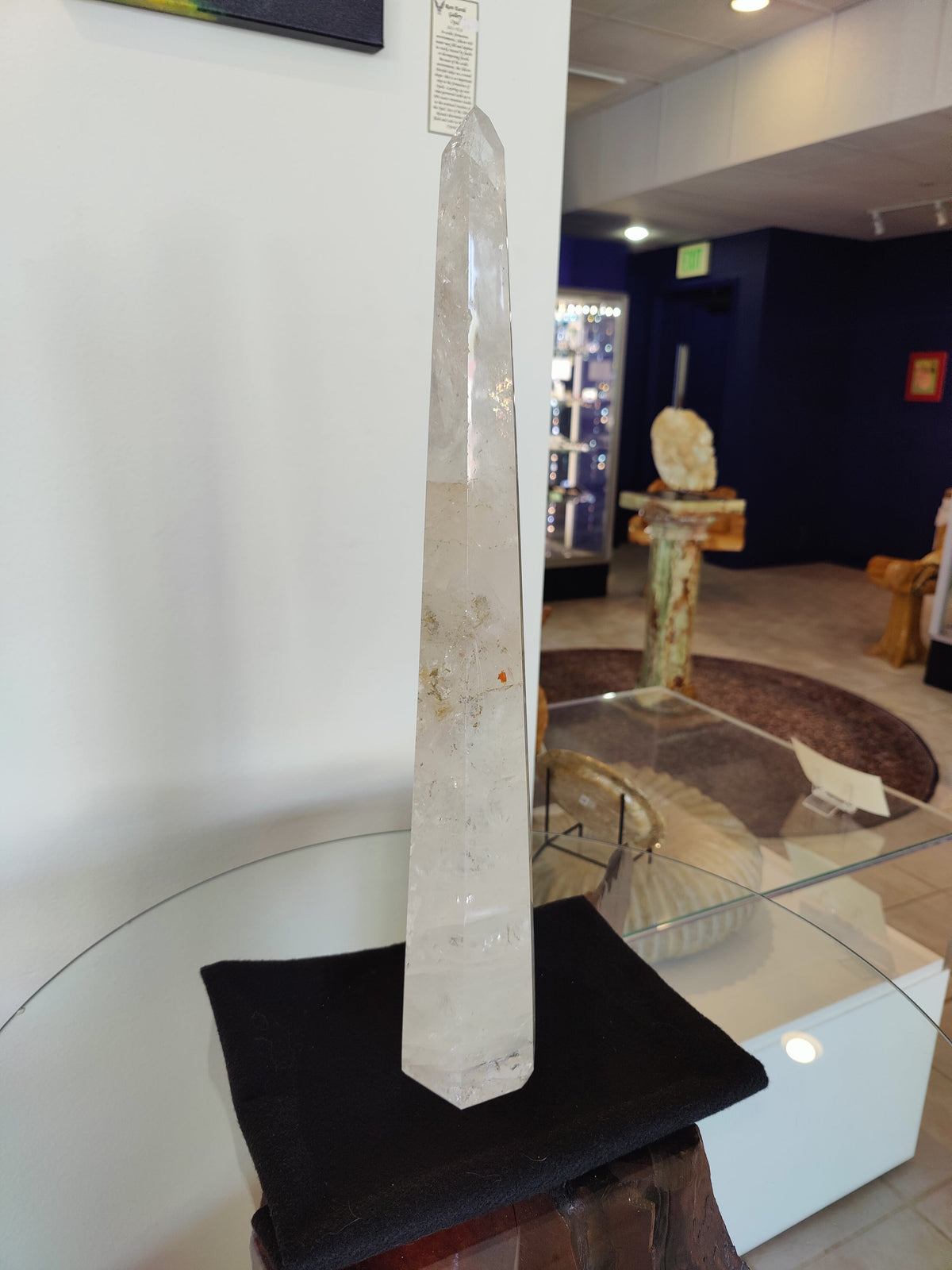 Tall, polished clear quartz obelisk with natural inclusions, standing on a black cloth against a white wall in a room with display cases and dark blue walls.