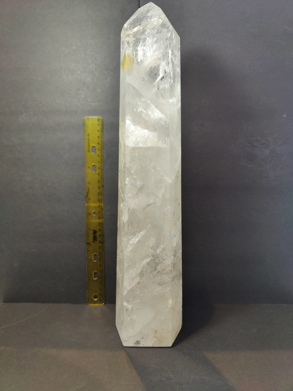 Tall polished clear quartz obelisk with natural internal fractures, standing upright next to a yellow measuring ruler for scale against a dark background.