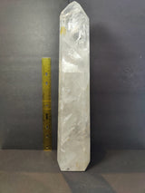 Tall polished clear quartz obelisk with natural internal fractures, standing upright next to a yellow measuring ruler for scale against a dark background.