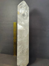 Tall, polished clear quartz obelisk with natural internal fractures, standing vertically next to a yellow ruler for scale on a dark background.