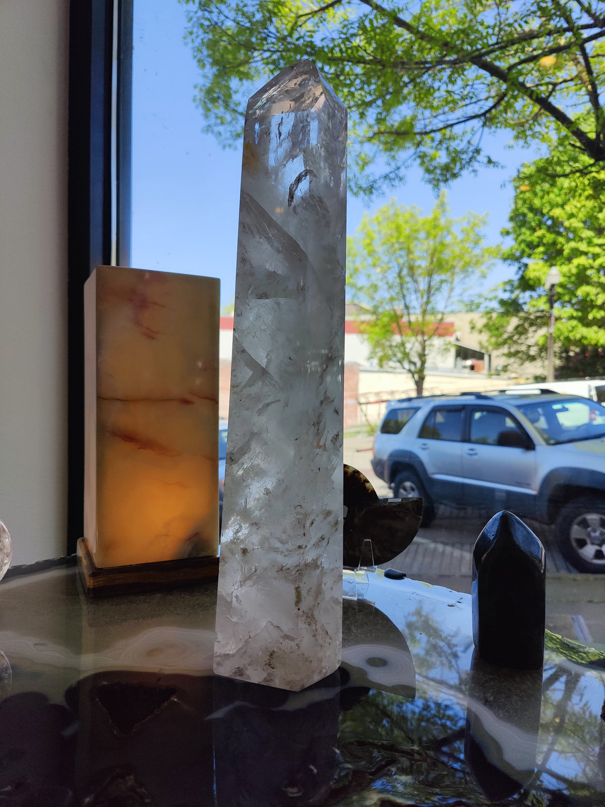 Tall clear quartz polished obelisk with faceted sides, standing on a glass surface near a window showing trees and a parked silver SUV outside, accompanied by smaller dark and amber stones.