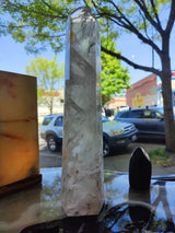 Tall, polished clear quartz obelisk with natural inclusions, standing on a reflective surface against an outdoor background with a tree and parked cars.