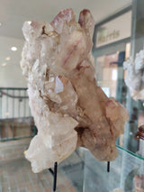 Translucent lithium quartz cluster with pointed crystal formations showing pinkish hues, displayed upright on black stands inside a glass case.