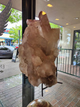 Large lithium quartz crystal cluster with translucent pale purple and beige hues, displayed on a black metal stand near a window showing an outdoor street scene.