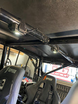 Interior roof of a UTV showing two bright dome lights attached with straps along a black metal roll cage above two black SPEED-branded bucket seats.