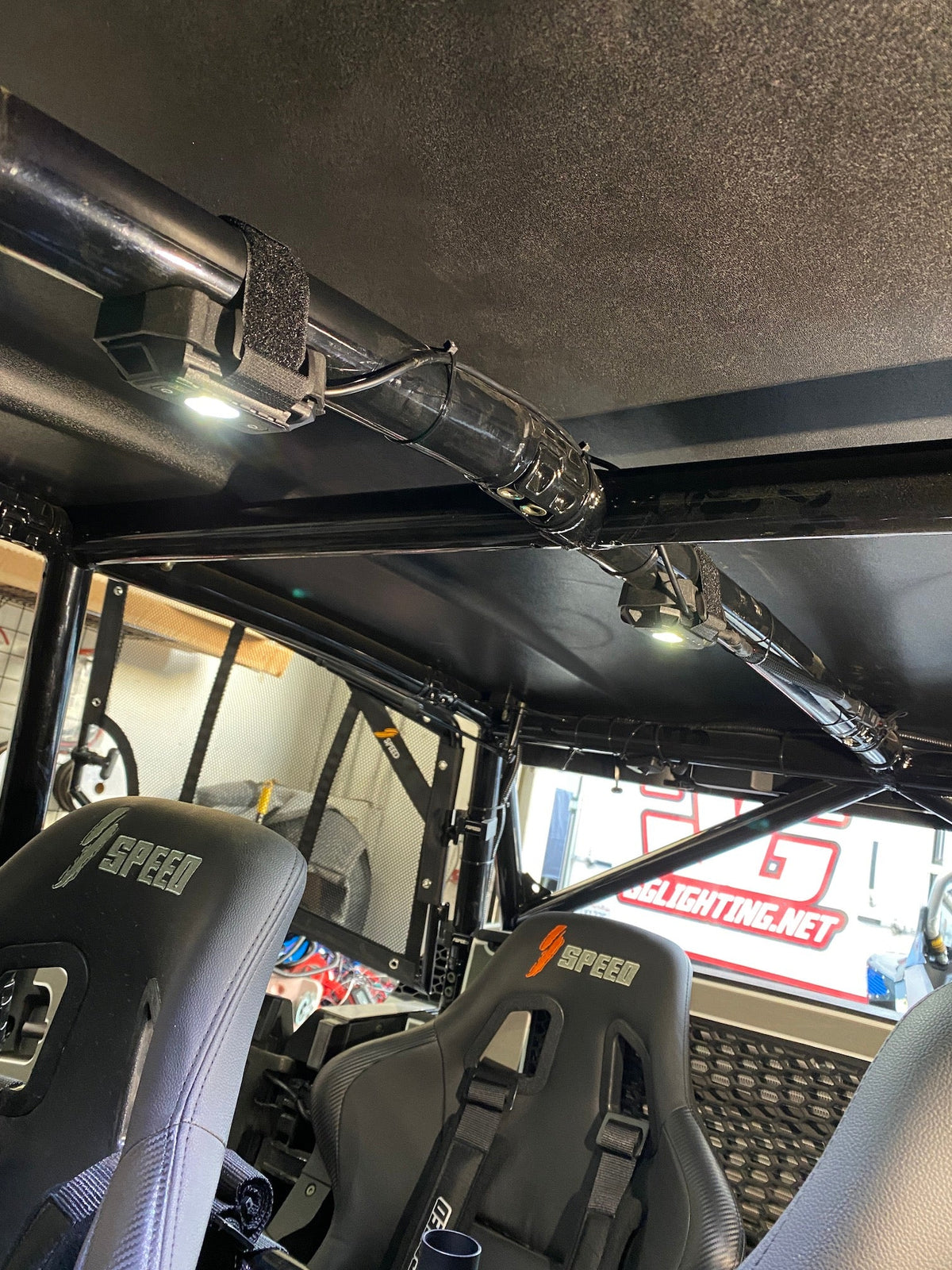 Interior roof of a UTV showing two bright dome lights attached with straps along a black metal roll cage above two black SPEED-branded bucket seats.