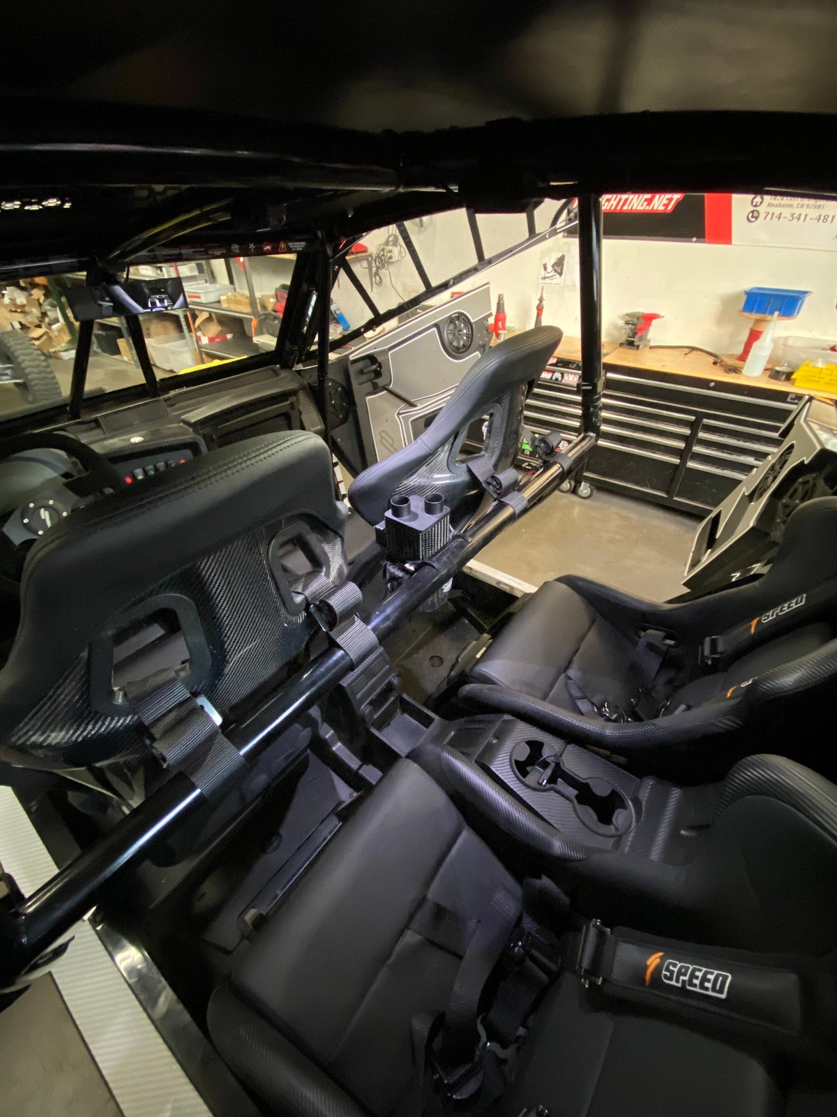 Interior of a UTV showing two black Speed brand bucket seats with harnesses and a roll cage, viewed from the back towards the dashboard and storage box.