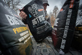 Man handling large black bags labeled Big Tine Platinum fortified deer blend outdoors on a truck bed with leafless trees in the background.