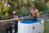 Muscular tattooed man in black shorts sitting on the edge of a white inflatable tub with a blue interior, outdoors next to green plants and a portable machine.