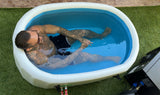 Man with tattoos sitting in a small white inflatable tub filled with water on green grass, viewed from above at an angle.