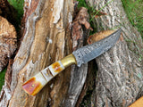 CUSTOM HANDMADE FORGED DAMASCUS STEEL HUNTING KNIFE with burnt bone and stag horn scales lying on a surface, showing layered Damascus steel pattern on blade and textured wood handle