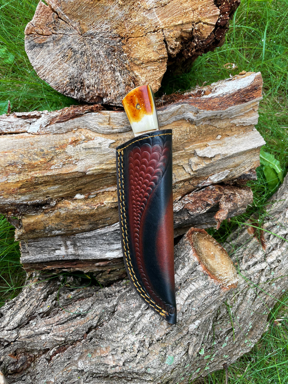 Forged Damascus steel hunting knife with burnt bone and stag horn handle scales in a dark carved leather sheath resting on stacked weathered wood logs on green grass.