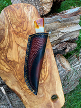 Hunting knife with burnt bone and stag horn handle partially sheathed in dark brown leather with scalloped pattern, resting on a light wood board outdoors near chopped wood.
