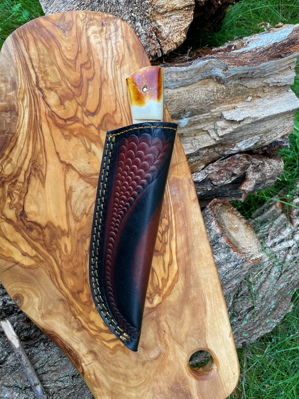 Hunting knife with burnt bone and stag horn handle partially sheathed in dark brown leather with scalloped pattern, resting on a light wood board outdoors near chopped wood.