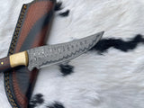 Hunting knife with patterned Damascus steel blade and light-colored bolster, resting on a brown leather sheath with black stitching on white and black spotted fur background.