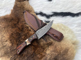 Damascus steel skinning knife with patterned blade, rosewood handle accented by a stag section, resting on a brown fur with a stitched leather sheath beside it