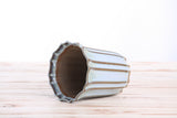 Ceramic bonsai pot with vertical ridges and scalloped edge, glazed in muted white and brown tones, shown lying on its side on a light wood surface.