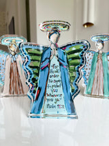 Translucent angel figurines with painted blue robes, green wings, and written Psalm 91:11 on the central figure, photographed on a reflective surface with blurred background figures.