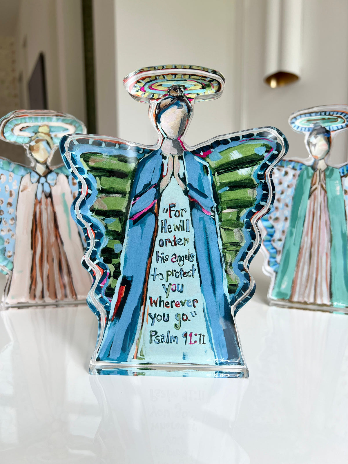 Translucent angel figurines with painted blue robes, green wings, and written Psalm 91:11 on the central figure, photographed on a reflective surface with blurred background figures.